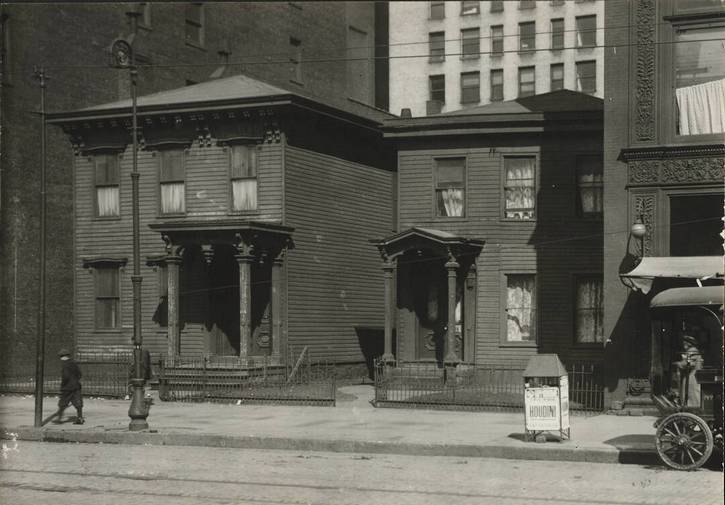 1910 rooming houses near the Rose Building, owned by livery operator John Corlett, who refused to sell the houses. Lower right, a sidewalk sign advertises a Houdini show at the Hippoddome.